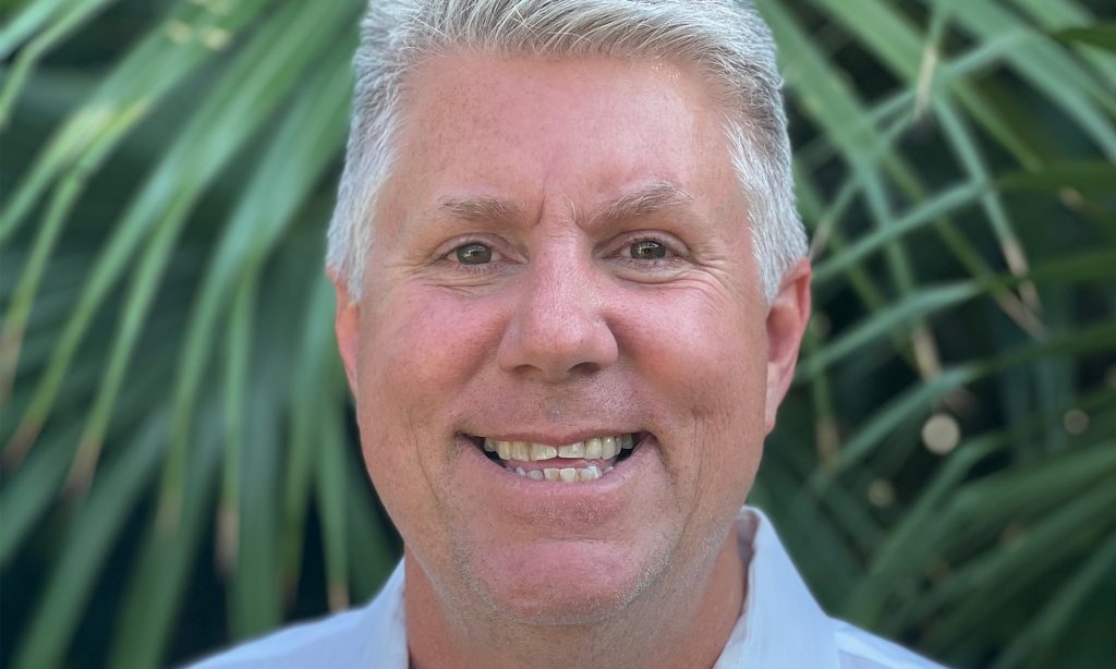 Man with gray hair smiling outside in front of trees.